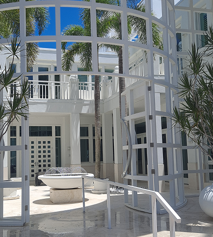 Modern architectural courtyard with white framed windows, palm trees, a circular fountain, and a ramp with a railing, leading to a bright blue sky 1